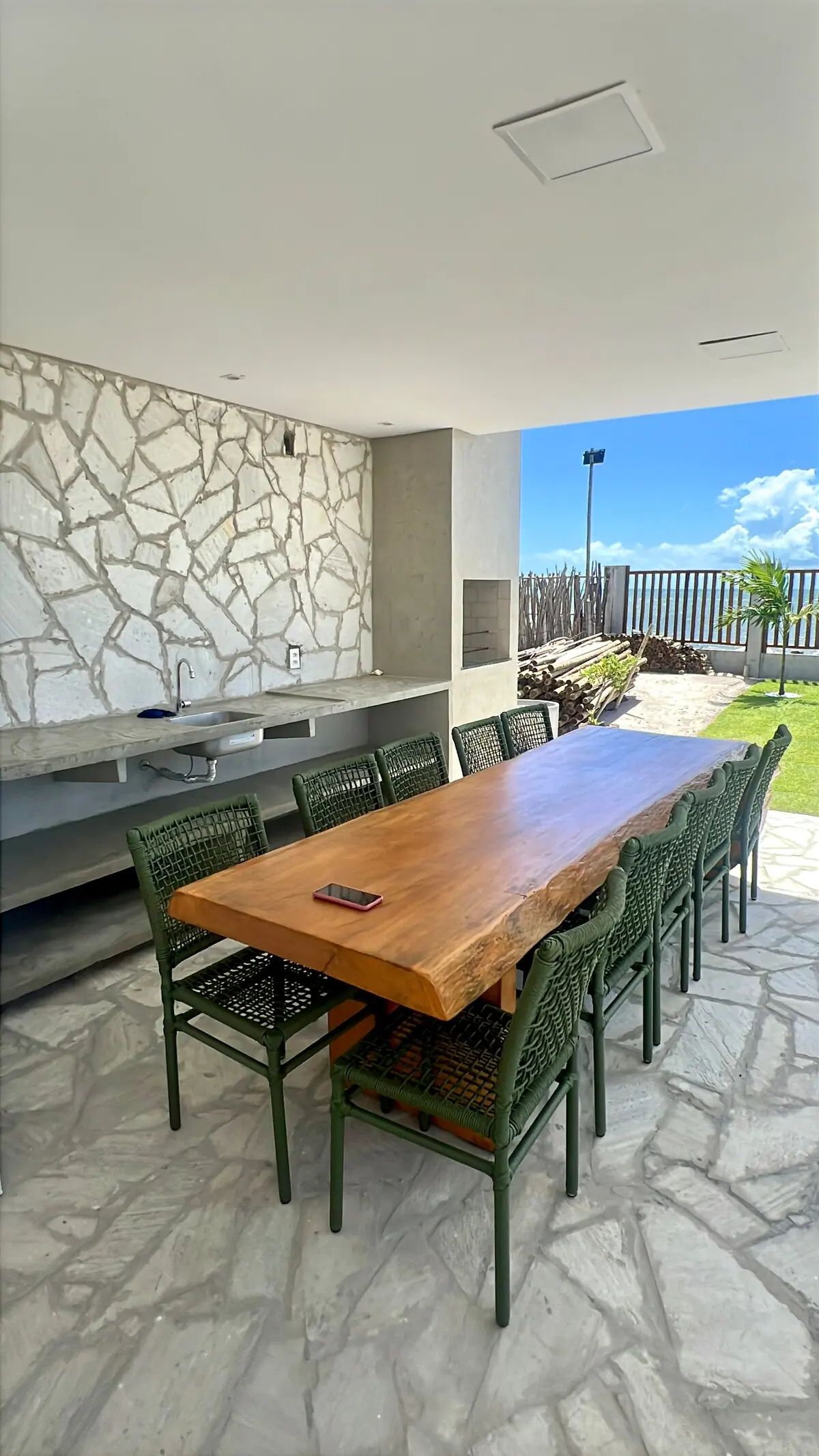 Área gourmet com mesa de madeira, cadeiras verdes e parede de pedra